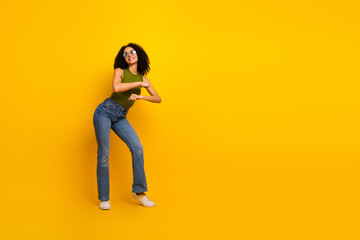 Cheerful young woman in casual green top and jeans posing playfully against a vibrant yellow...