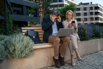 Stressed businessman and businesswoman reading bad news on laptop outdoors in business district,...