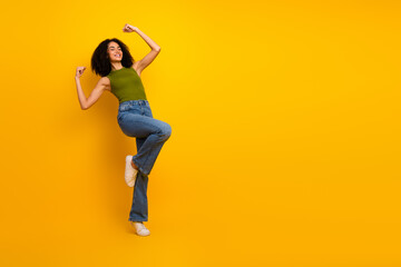 Attractive young woman in casual green top and jeans dancing on vibrant yellow background with...