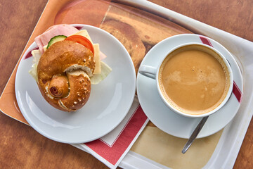 A delicious breakfast served on a tray, featuring a fresh pretzel roll sandwich filled with ham, cheese, lettuce, cucumber, and tomato on a white plate.