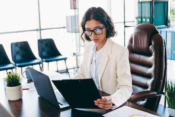 Confident businesswoman reviewing documents in a modern office workspace with sunlight filtering...