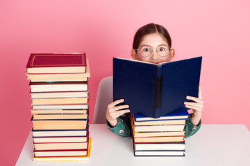 Adorable young girl reading a book surrounded by stacks of book against a pink background...