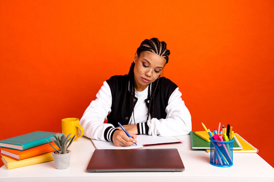 Young female student studying with determination surrounded by colorful objects and school supplies on a vibrant orange background