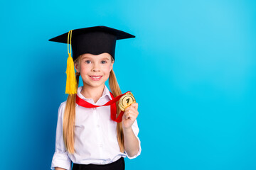 Smiling young schoolgirl with graduation cap holding gold medal on bright blue background...