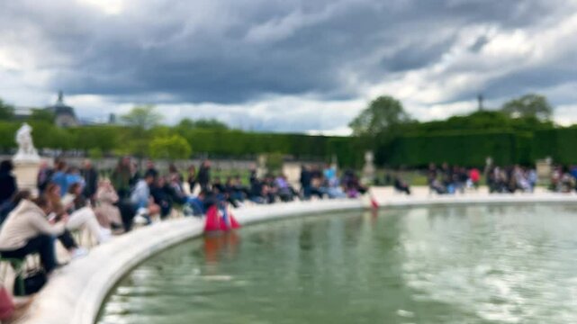 People sit and relax around the Grand bassin rond (Large round pool), small models of sailboats float on the water in Jardin des Tuileries, 1st arrondissement. Out of focus. Paris, France.