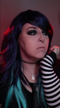 Alternative girl with dark makeup, striped sleeves, and facial piercings posing indoors under moody red and purple lighting.
