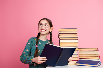 Cheerful young girl with braids reading a book amidst a stack of book against a vibrant pink...