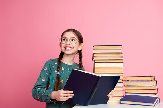 Cheerful young girl with braids reading a book amidst a stack of book against a vibrant pink background