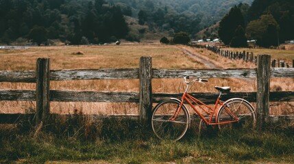 Fototapeta premium Vintage orange bicycle resting against rustic wooden fence in serene countryside landscape surrounded by lush green mountains