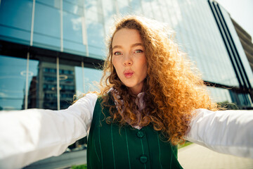 Young professional woman with curly red hair posing outdoors near modern urban building on a sunny...