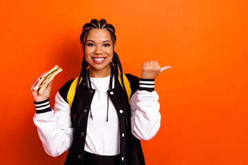 Cheerful student holding a sandwich against an orange background, wearing casual varsity jacket and...