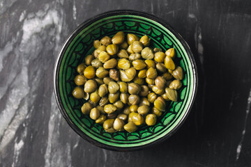 Pickled capers in green bowl. Salty preserved flower buds on dark marble surface. Mediterranean...