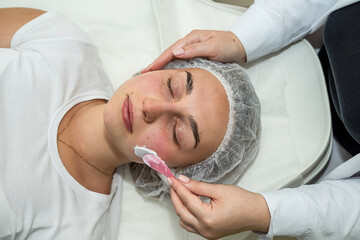 Beautician applies cleansing and moisturizing white cream with help of brush on client's face