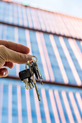 Hand Holding Keys in Front of Modern Glass Office Building. A close-up image of a hand holding a set of metallic keys against the background of a sleek glass skyscraper