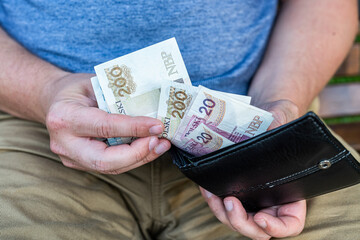 Man Holding Polish Zloty Banknotes in Wallet – Personal Finance Concept. Close-up of a man sitting on a bench and holding a black leather wallet with visible Polish zloty banknotes.