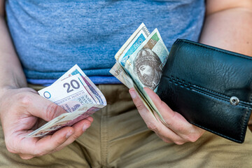 Man Holding Polish Zloty Banknotes in Wallet – Personal Finance Concept. Close-up of a man sitting on a bench and holding a black leather wallet with visible Polish zloty banknotes.