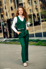 Confident young businesswoman in elegant green attire walking outdoors in a city setting on a sunny...