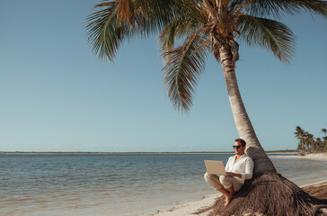 A man in shorts and sunglasses, wearing a hat, is sitting on the trunk of an old palm tree by the water