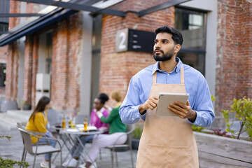 A Hindu male waiter holds a tablet, looking away from the camera at an outdoor restaurant. People sit at a table in the background.