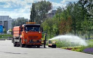 A worker waters flowers in a park with a hose. The water comes from a watering truck standing on the sidewalk. Improvement of the city park.
