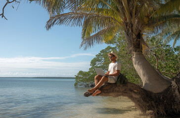 A man in shorts and sunglasses, wearing a hat, is sitting on the trunk of an old palm tree by the water