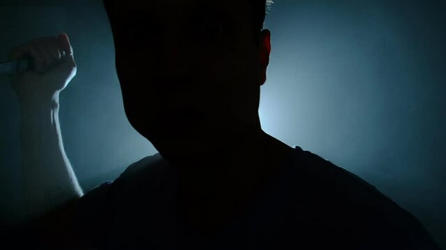 Silhouette of man approaching with knife in hand under blue backlight, cinematic danger and suspense concept