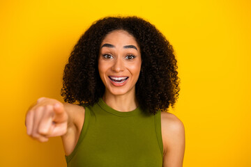 Cheerful young woman with curly hair pointing excitedly in a bright yellow background, wearing a...