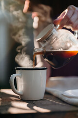 A woman’s hands gently pour fresh coffee into a white cup on a rustic wooden table in a cozy garden. Soft morning light and cottagecore atmosphere complete the scene.