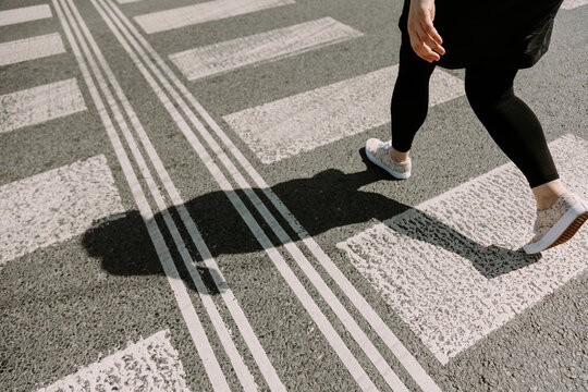 A woman walking briskly across a sunlit crosswalk with a sharp shadow falling on the pavement. Ideal for themes of lifestyle, urban movement, freedom, or motivation