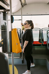 Stylish woman wearing a black outfit stands by the exit in a city bus, holding a pole and wearing sunglasses. Great for depicting daily urban commute, public transport, and modern lifestyles.
