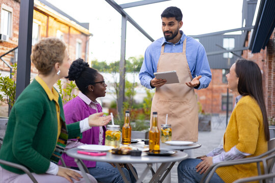 A waiter taking orders from a group of friends at an outdoor cafe, using a tablet device. - Powered by Adobe
