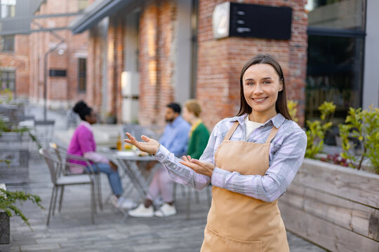 Smiling waitress welcomes customers to an outdoor restaurant, gesturing towards the seating area with other patrons.