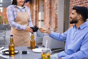 A smiling female waiter presents a payment terminal to a male customer seated at an outdoor cafe...