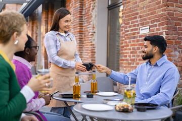 A female waiter is at a table and is asking a customer to pay the bill using a portable payment terminal. Other customers are present.