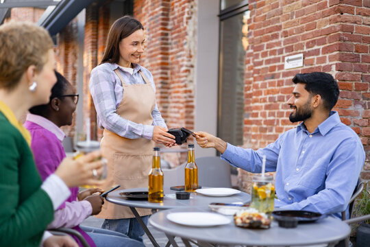 A female waiter is at a table and is asking a customer to pay the bill using a portable payment terminal. Other customers are present. - Powered by Adobe