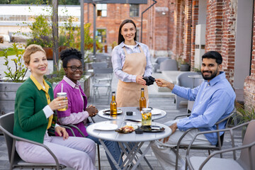 A female waiter in an apron serves customers at an outdoor restaurant, accepting payment via a...