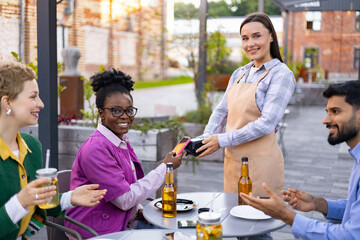 A female waiter in an apron uses a portable terminal to process a payment at an outdoor restaurant,...