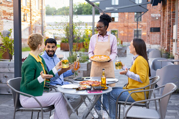 A waitress serves pizza and drinks to a group of friends at an outdoor restaurant. They are enjoying their time.