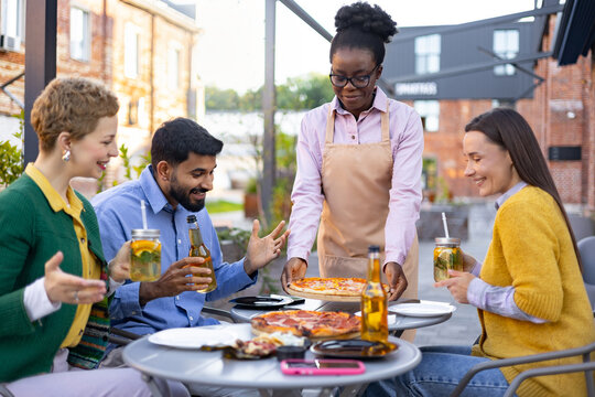 A friendly waitress serves pizza to a group of friends at an outdoor restaurant, enjoying drinks.