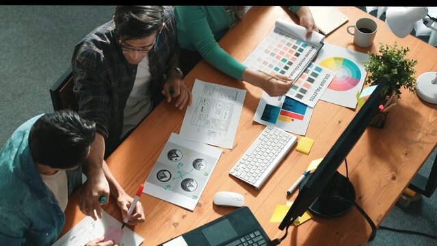 Top view of manager writing idea while team standing and giving comment. Professional designer design logo while looking at color palette and writing idea down in paper with color marker. Symposium.