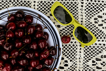 Cherries and green stylish sunglasses create a vibrant summer still life scene. A plate filled with fresh, ripe cherries next to a pair of yellow sunglasses on a lace-patterned surface Summertime vibe