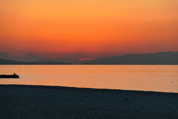 Sun dips below distant mountains across the sea, painting the sky in intense shades of orange and red during a breathtaking sunset viewed from a quiet, deserted beach, creating a dramatic landscape.