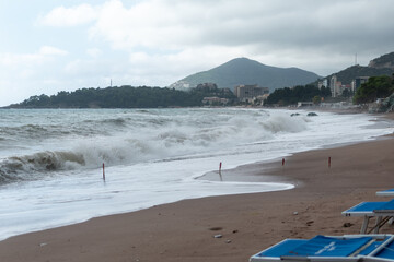 Stormy beach scene with large waves crashing onto the sand, backed by distant buildings and a...