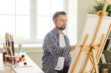 Young 25s European male artist painting on easel at home studio or workshop, looks focused on work...