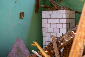 Interior of a ruined house with a white tiled stove, green wall, and collapsed wooden debris
