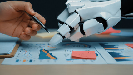 A human hand and a robotic hand work together on business charts, analyzing data trends and...