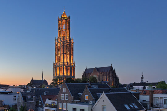 Experience the enchanting glow of a historic European city at dusk, highlighting the magnificent illuminated Dom Tower "Domtoren" in Utrecht rising above charming rooftops.