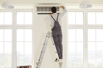 Service worker wearing overalls, standing on ladder, repairing air conditioner, checking if there...