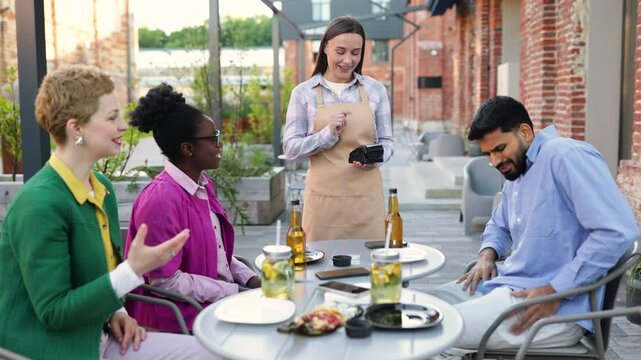A female waiter is at a table and is asking a customer to pay the bill using a portable payment terminal. Other customers are present.