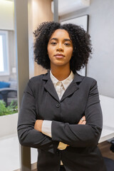 Confident young Black businesswoman with curly hair standing with arms crossed in a modern office,...
